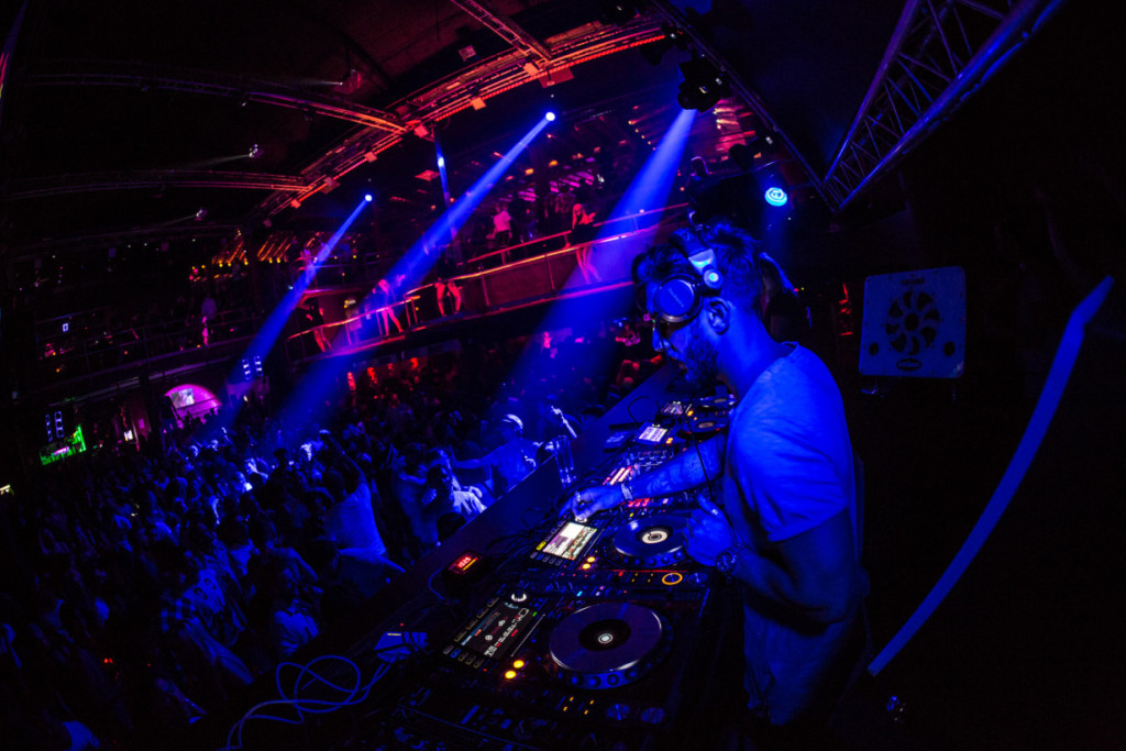 DJ mixing in a dark club washed in blue light while a packed dance floor moves below