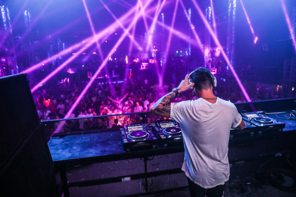 DJ performing behind the decks in a dark club lit by magenta laser beams, crowd dancing in front of the booth